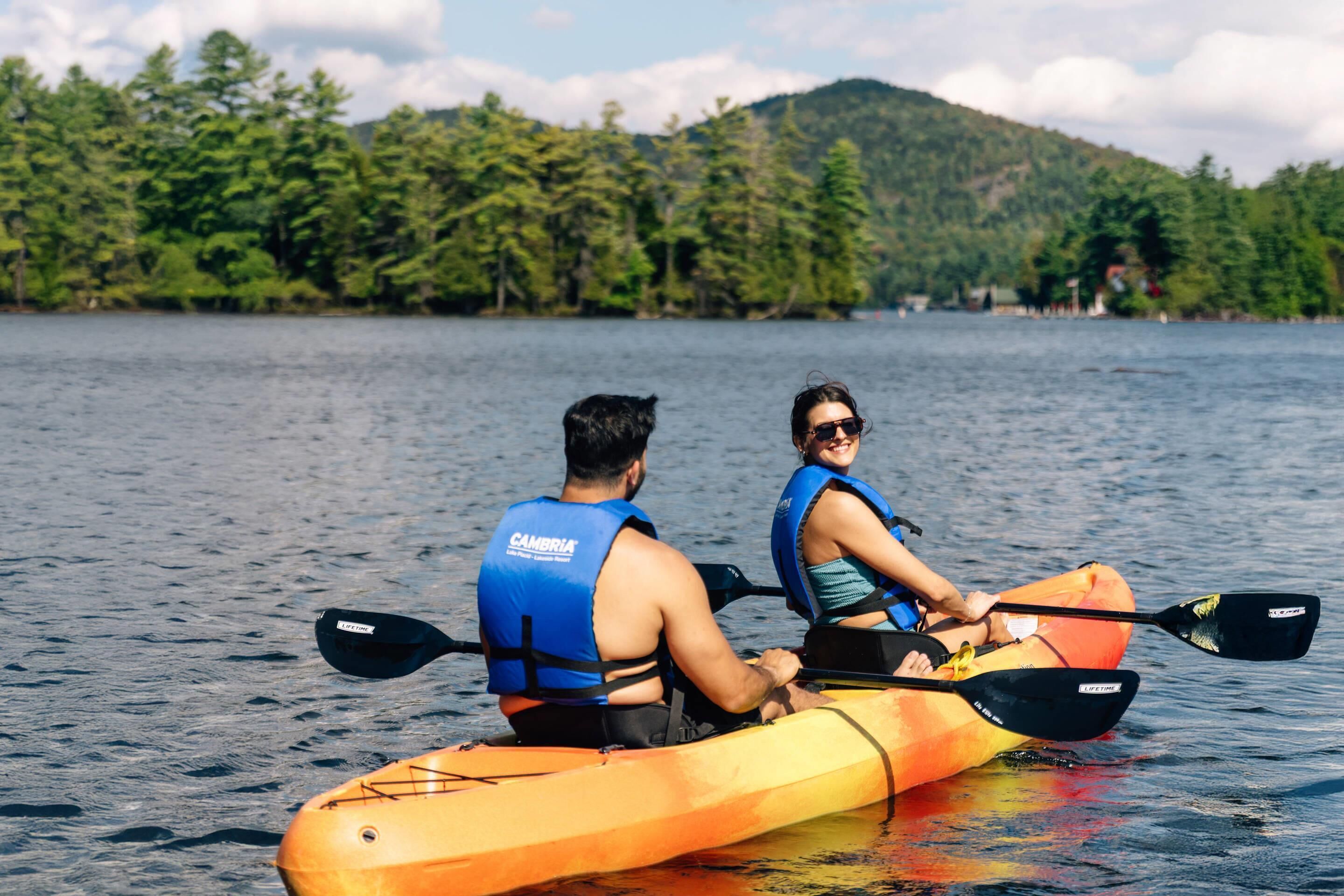 Two people kayaking on a lake with a forested shoreline in the background.