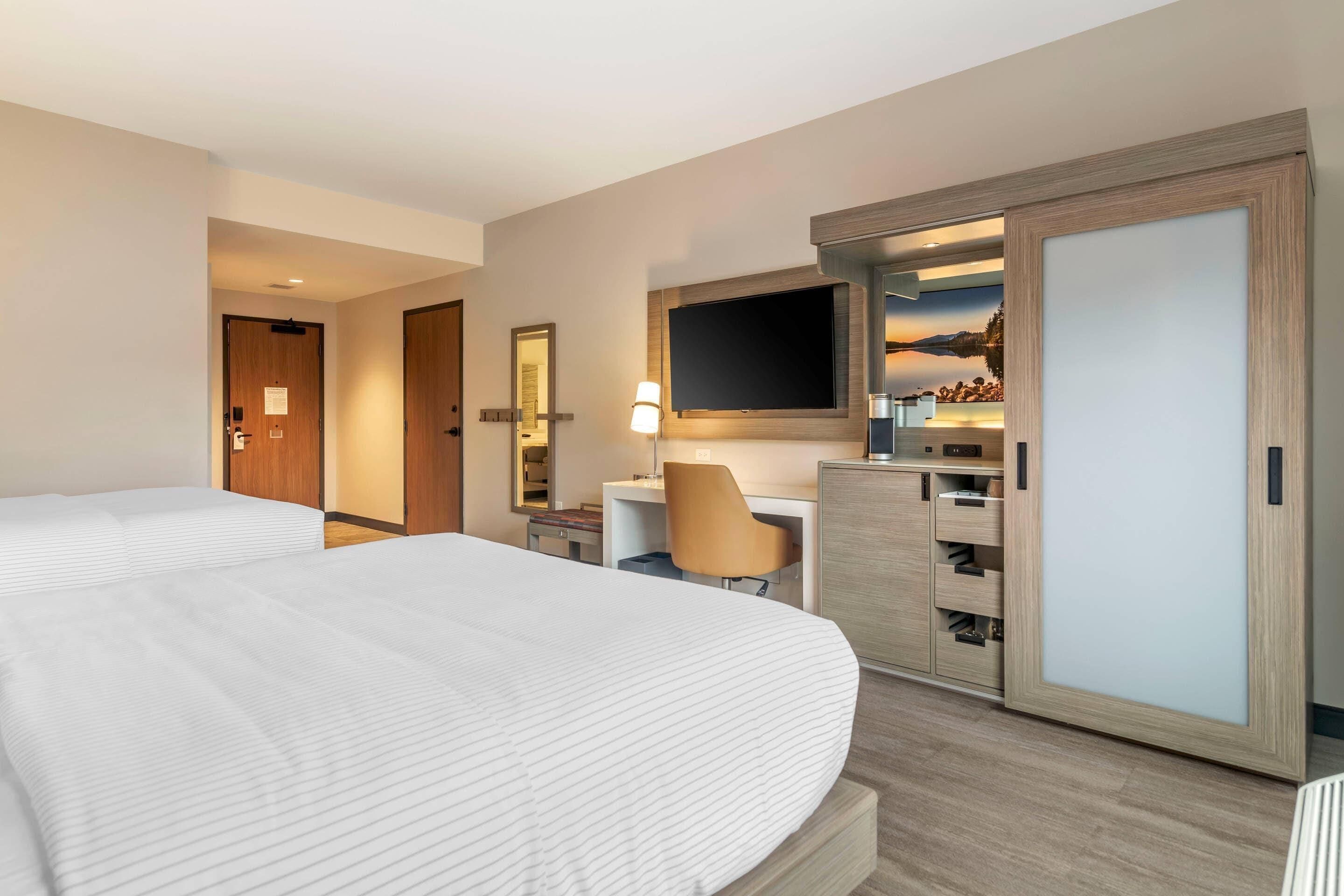 A hotel room with a large bed, a desk with a chair, a TV mounted above the desk, and a wardrobe.