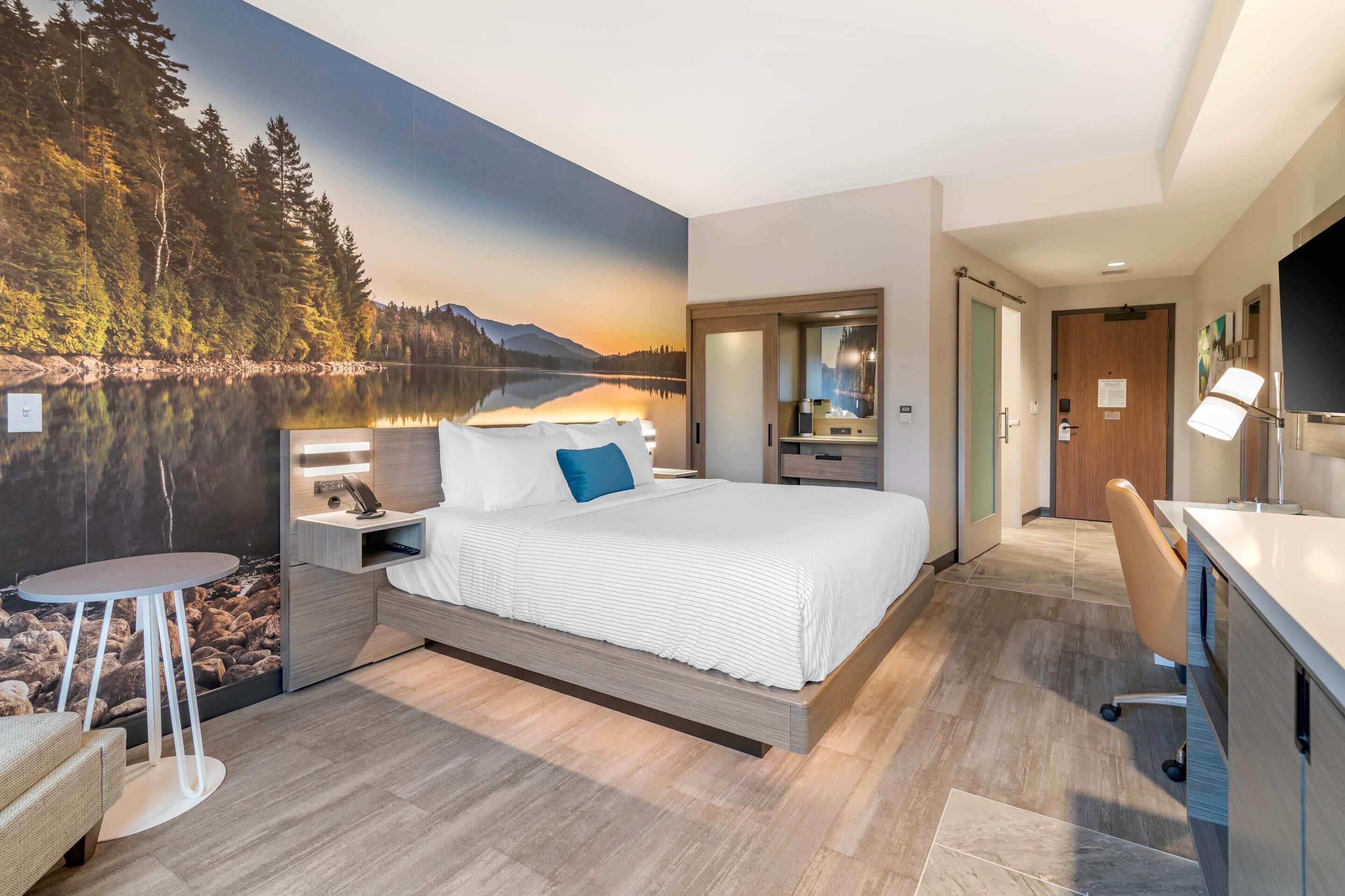 A modern hotel room with a large bed, a desk, and a view of a lake through a mural.