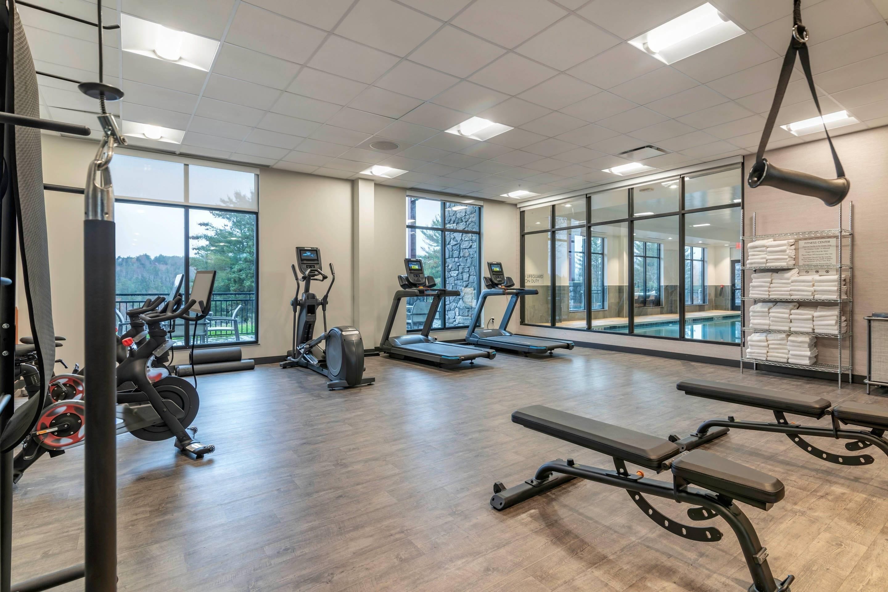 A gym with cardio machines, benches, and a pool visible through the windows.