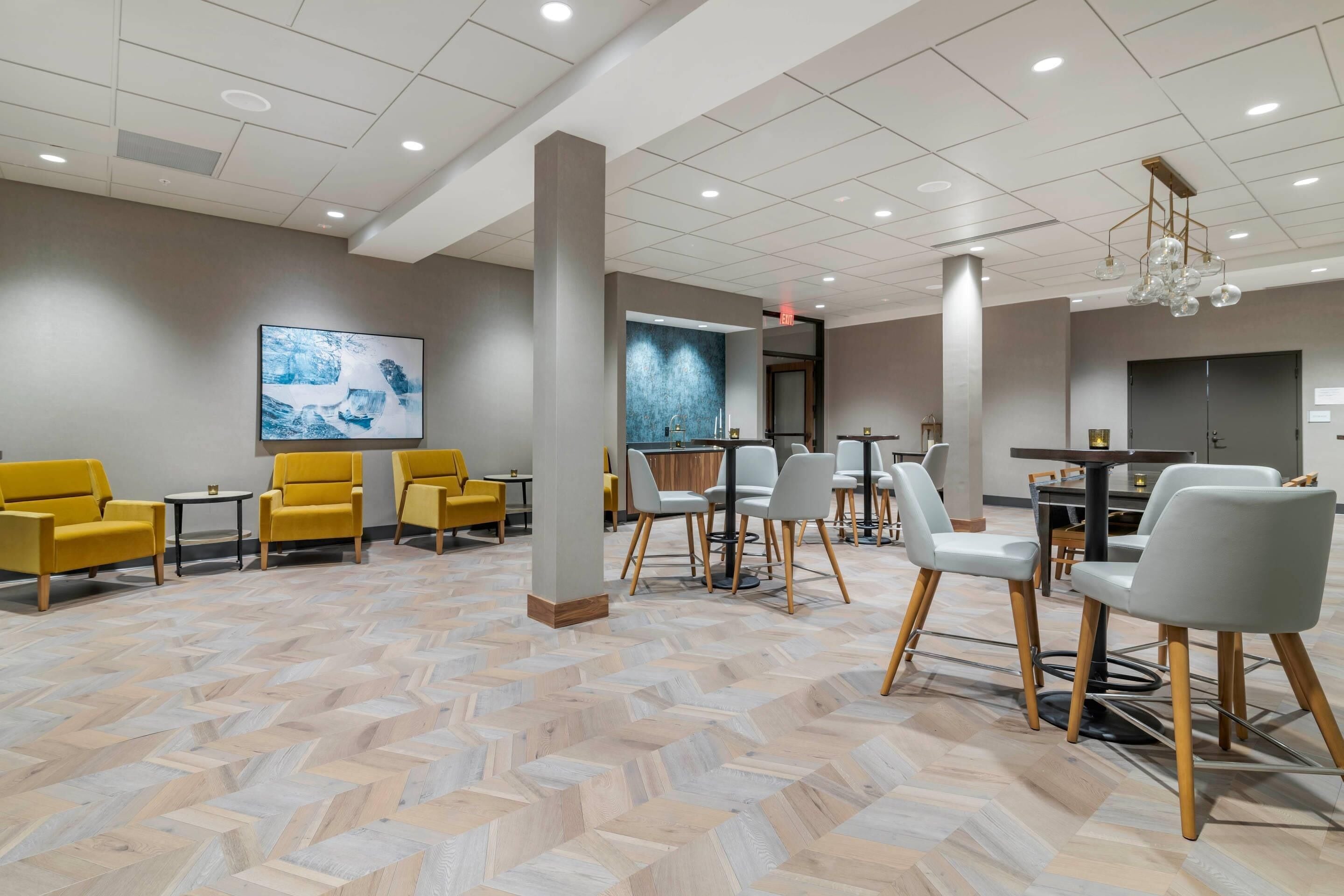 A modern hotel lobby with a herringbone-patterned floor, yellow and gray seating, and a contemporary chandelier.