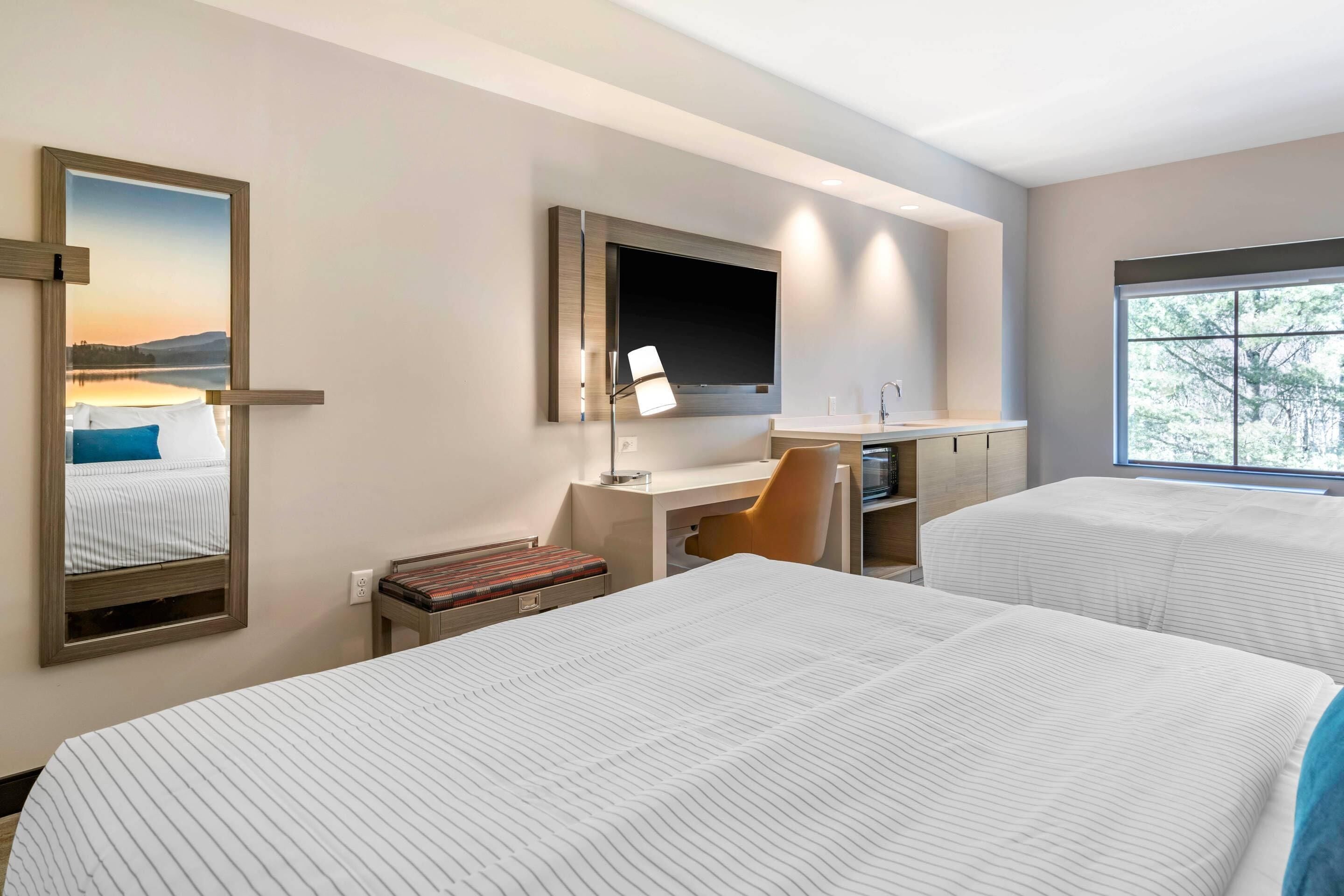 A hotel room with two beds, a desk with a chair, a TV mounted on the wall, and a window providing a view of trees.