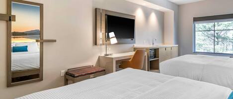 A hotel room with two beds, a desk with a chair, a TV mounted on the wall, and a window providing a view of trees.