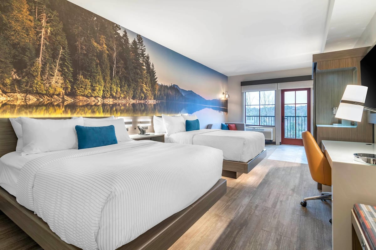 A hotel room with two beds, a desk, a chair, and a large forest mural on the wall.