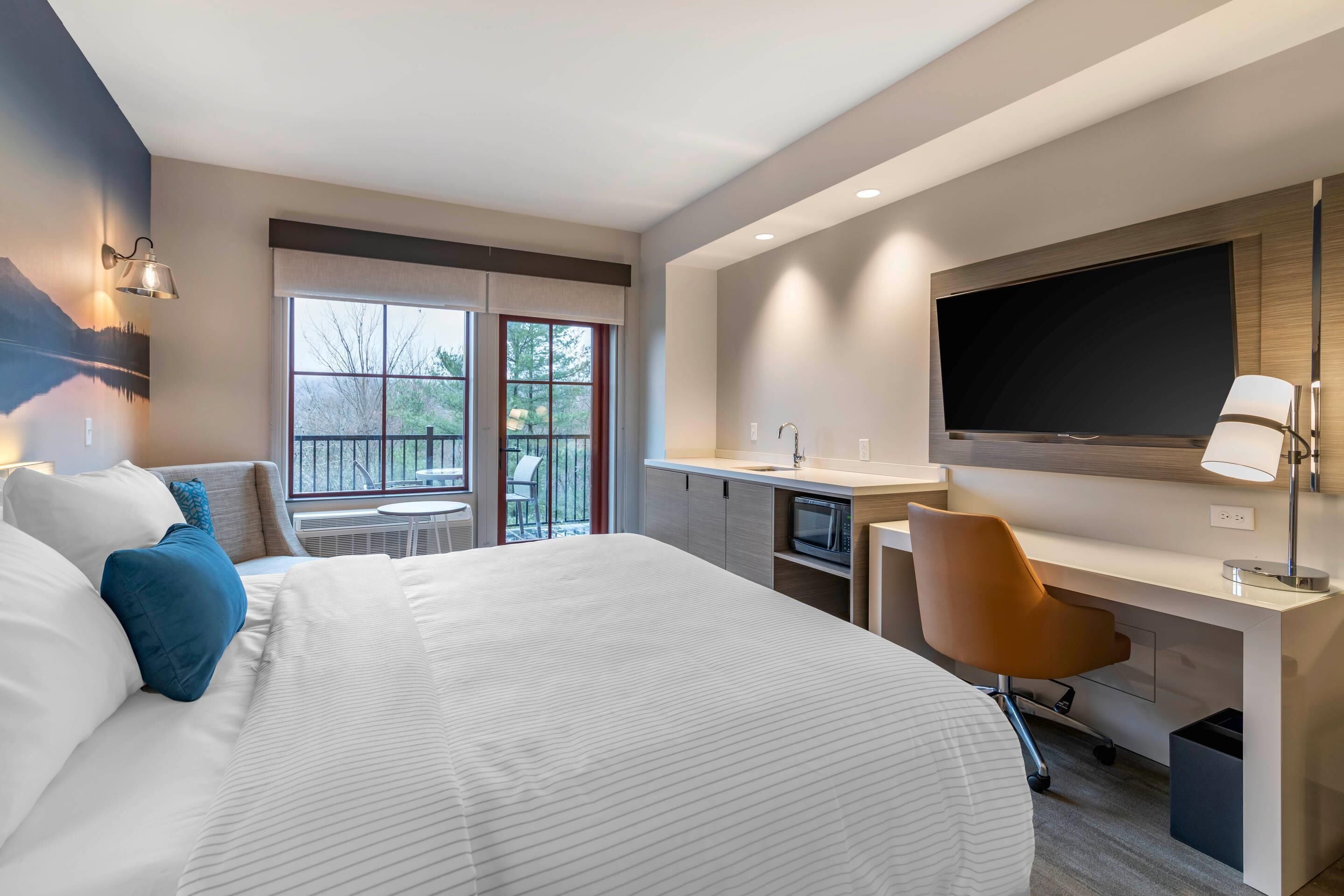 A modern hotel room with a large bed, a desk with a TV, and a balcony with a view of trees.