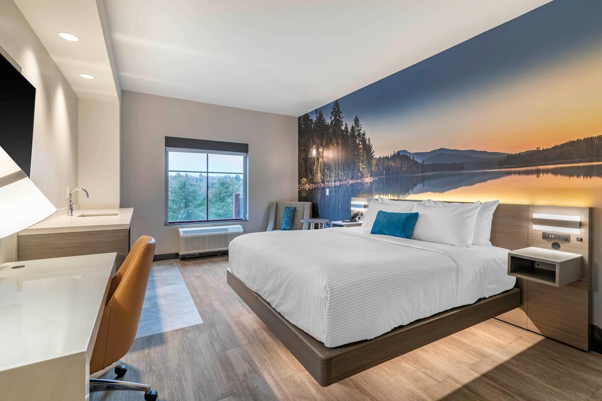 A modern hotel room with a large bed, a desk, a chair, a nightstand, a lamp, and a window with a view of a lake and mountains.