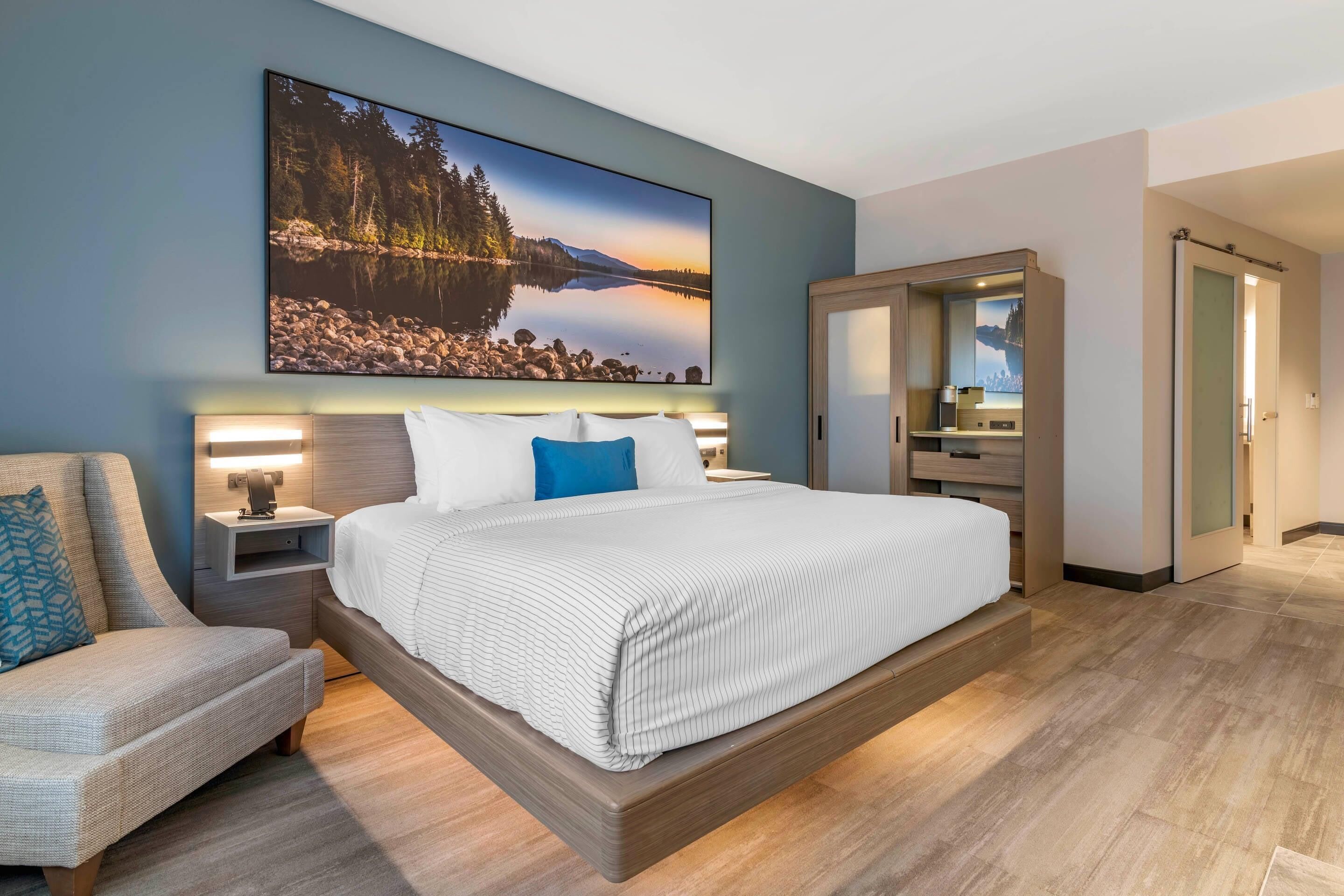 A modern hotel room with a large bed, a seating area, a bathroom, and a view of a lake.