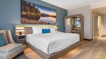 A modern hotel room with a large bed, a seating area, a bathroom, and a view of a lake.