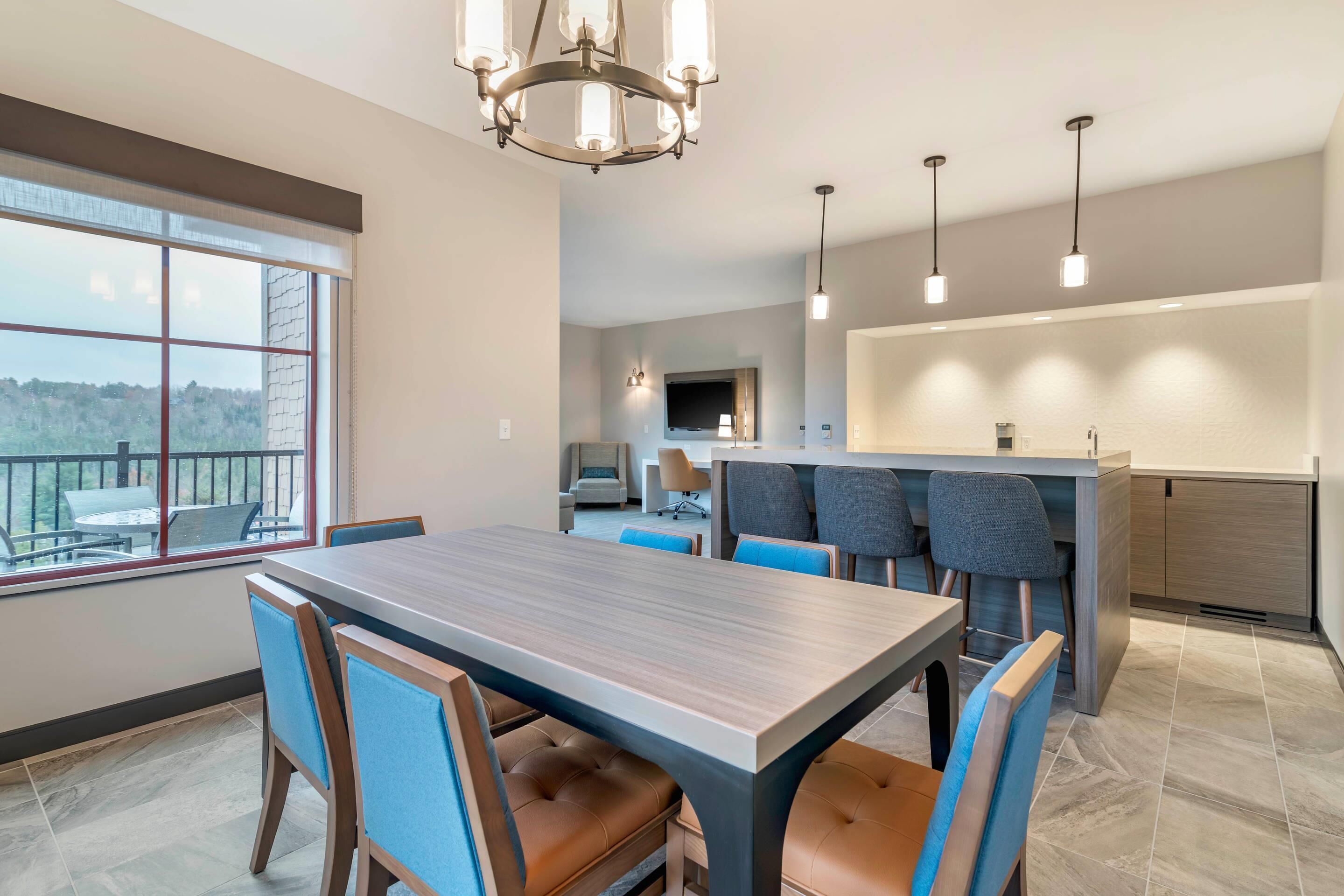 A modern dining area with a large table and chairs, a chandelier, and a view through large windows.