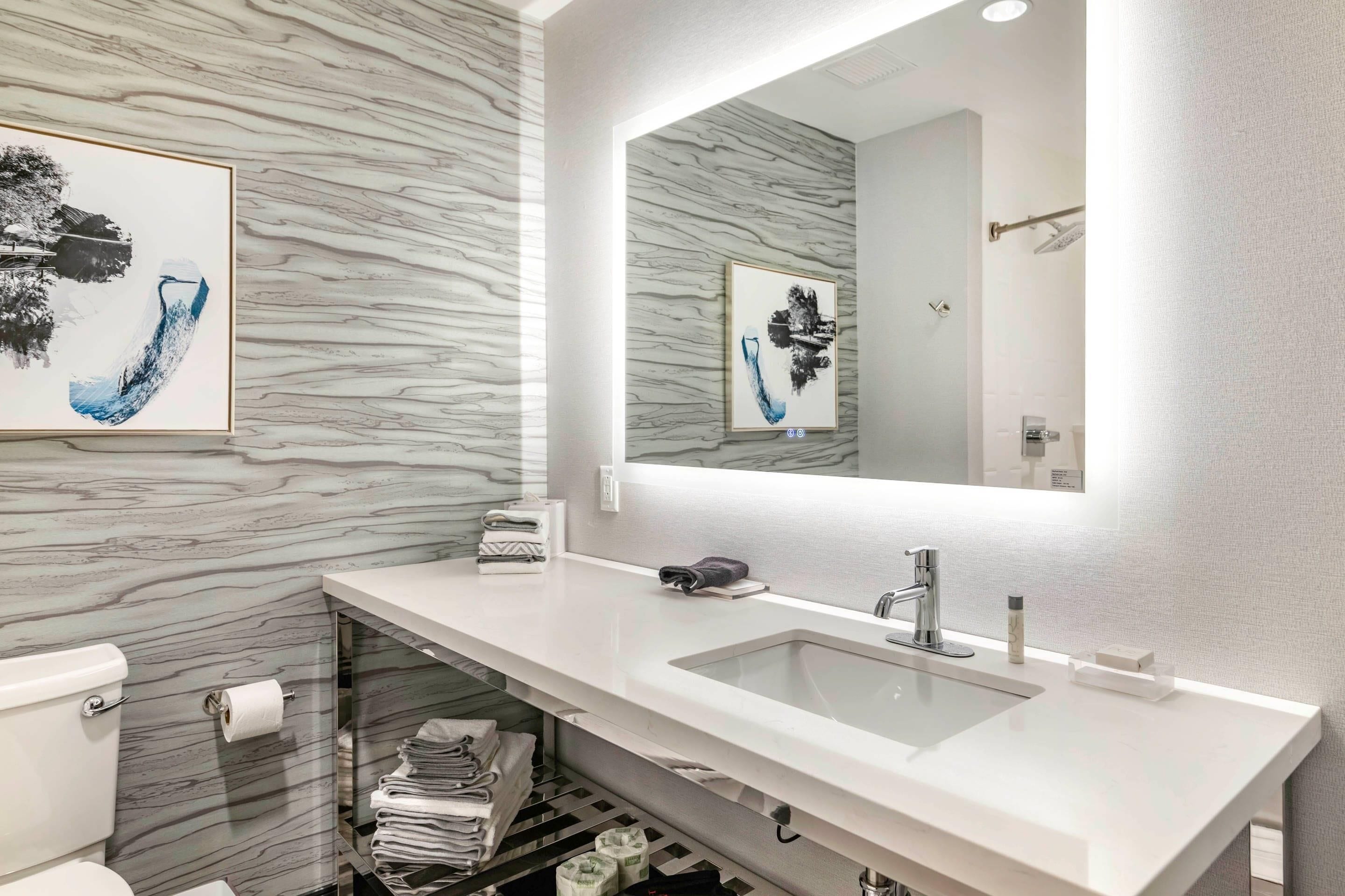 A modern bathroom with a marble backsplash, a large mirror, a white sink, and neatly arranged towels.