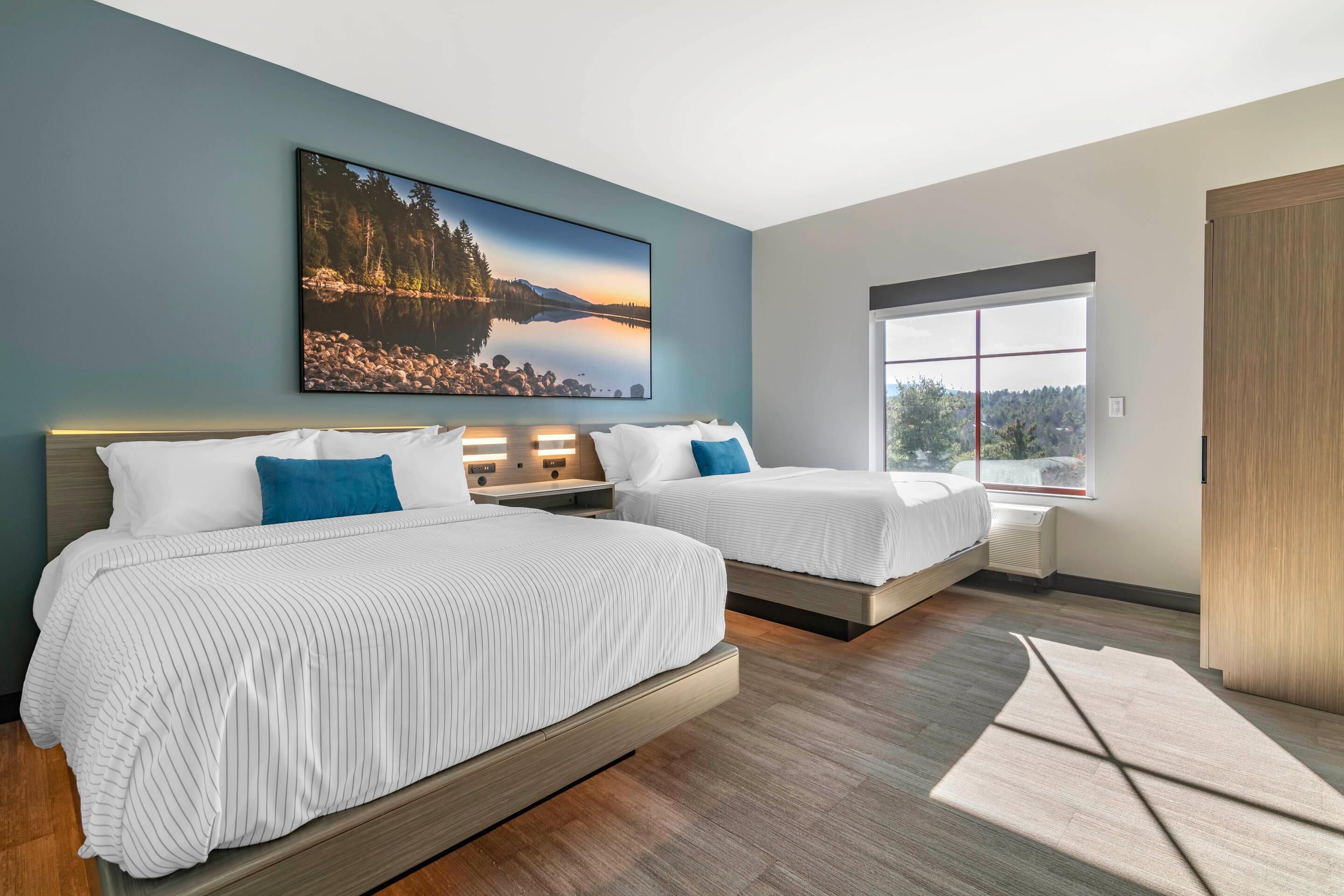 A modern bedroom with two beds, a large window, and a painting of a lake.
