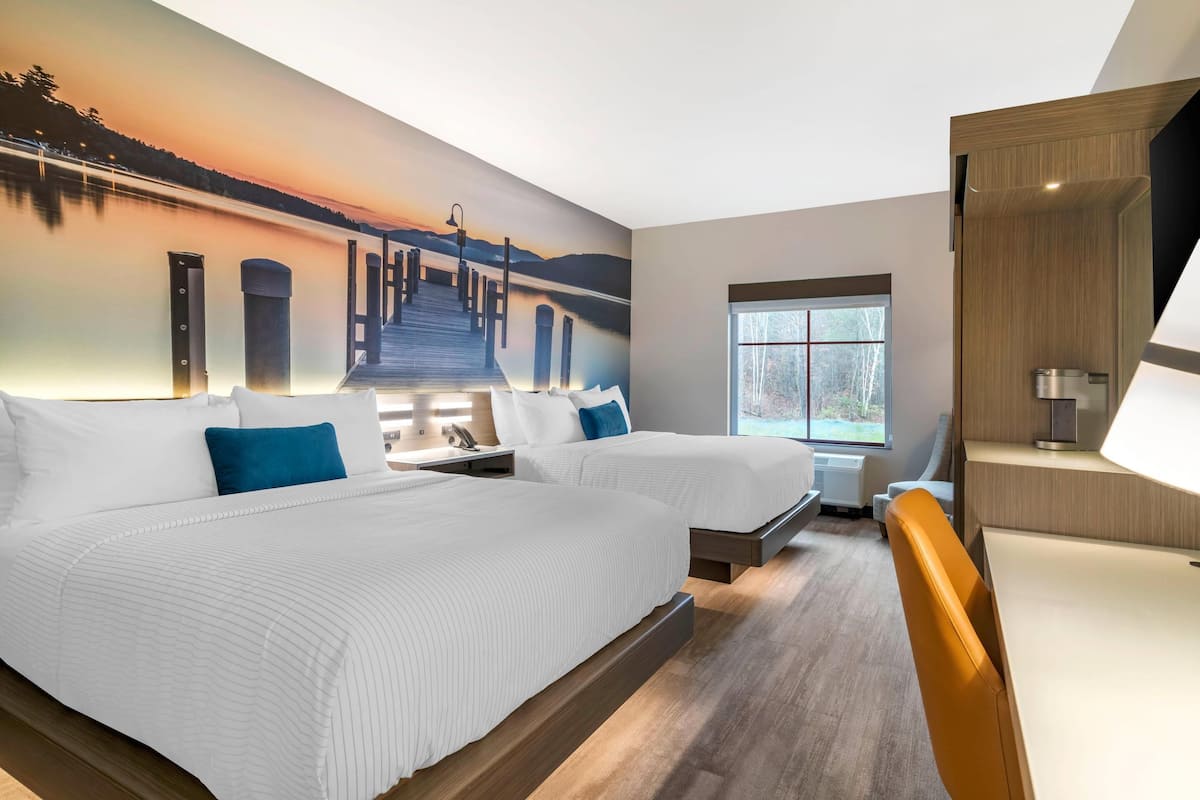 A hotel room with two beds, a large mural of a sunset over a pier, a window with a view of trees, and a modern desk with a lamp.