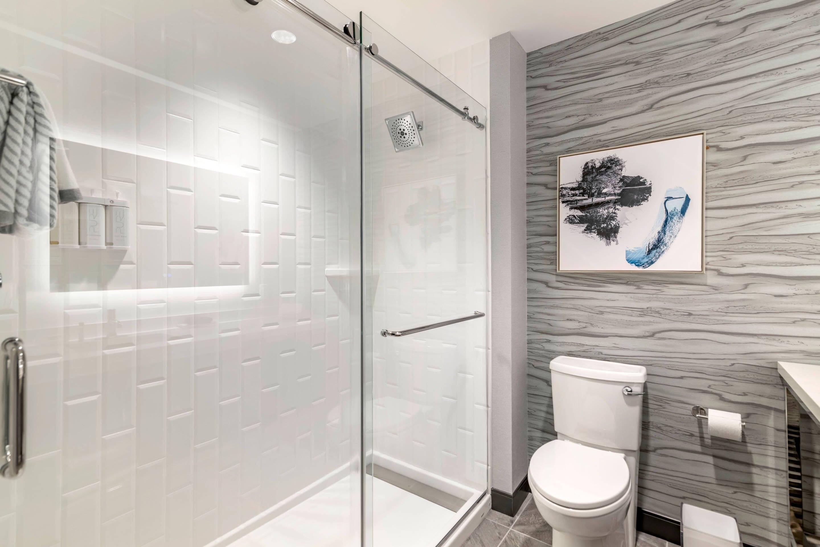 A modern bathroom with a glass-enclosed shower, white tiled walls, a marble-patterned accent wall, and a framed artwork above the toilet.