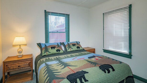 A bedroom with a bed, two windows with blinds, a nightstand with a lamp, and a ceiling fan. - Nana's Cottage Retreat (Big Bear City)