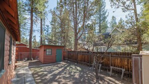 A backyard with a wooden cabin, a small shed, and a fenced area with tall trees. - Nana's Cottage Retreat (Big Bear City)