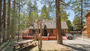 A cabin with a wooden deck, picnic tables, and a fenced area in a forest setting. - Nana's Cottage Retreat (Big Bear City)