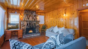 A cozy cabin interior with a stone fireplace, wooden walls, a sofa, and a desk with a television. - Nana's Cottage Retreat (Big Bear City)