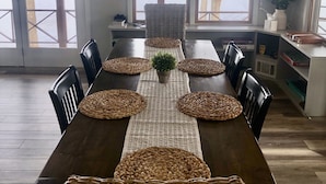 Un espace repas avec une longue table, des sets de table en fibres tressées et des chaises. De grandes fenêtres offrent une vue sur une étendue d’eau.