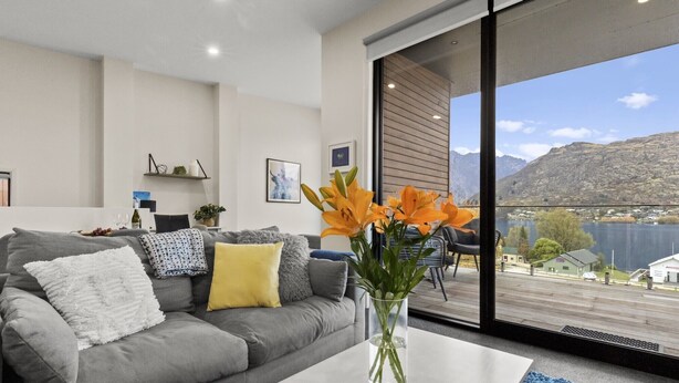 Ein modernes Wohnzimmer mit einer grauen Couch, einem weißen Couchtisch und einer Vase mit orangefarbenen Blumen. Ein großes Fenster bietet Blick auf einen See und Berge.