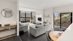 Un salon moderne avec un canapé gris, une table à manger avec un verre à vin, et une grande fenêtre offrant une vue panoramique.