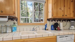 A kitchen with wooden cabinets, a double sink, and a window with a view of trees. - Cozy Fox Cabin (Big Bear Lake)