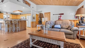 A spacious living area with a wooden ceiling, a kitchen in the background, and a cozy seating area with a sofa and a coffee table. - Cozy Fox Cabin (Big Bear Lake)