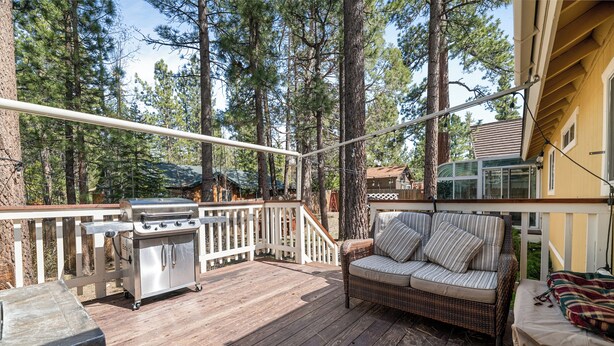 A wooden deck with a grill, a wicker sofa, and a railing overlooking a forest. - Cozy Fox Cabin (Big Bear Lake)
