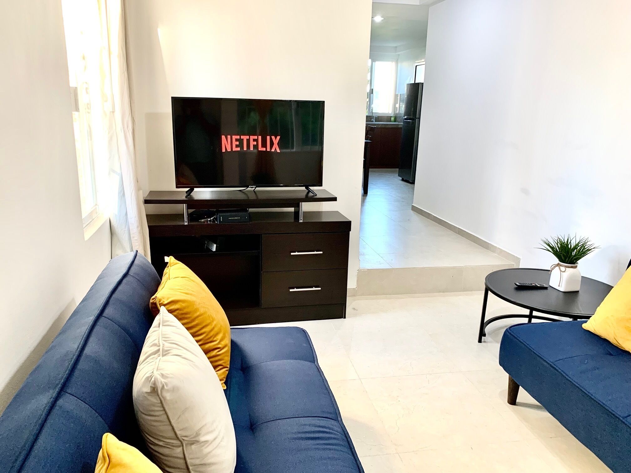A modern living room with a flat-screen TV on a black stand, a sofa with yellow and white cushions, and a small table with a plant.