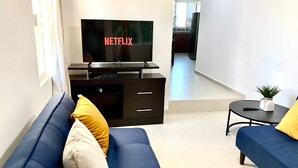Una sala de estar moderna con un televisor de pantalla plana sobre un soporte negro, un sofá con cojines amarillos y blancos, y una mesita con una planta.