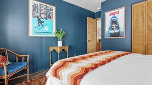A bedroom with a bed, a chair, a small table with a plant, and two posters on the wall.