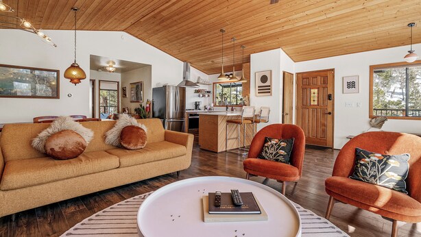 A spacious living room with a wooden ceiling, a sofa, two armchairs, a coffee table, and a kitchen area in the background.