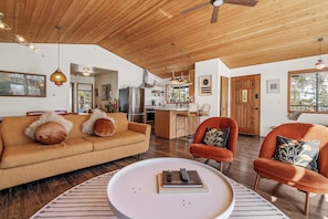 Uma sala de estar ampla com teto de madeira, um sofá, duas poltronas, uma mesa de centro e uma área de cozinha ao fundo.