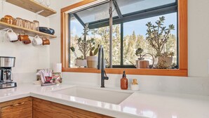 A kitchen with a white countertop, wooden cabinets, a stainless steel sink, and a window with a view of trees.