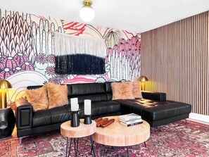 Un salon moderne avec un canapé en cuir noir, deux petites tables d’appoint rondes en bois, un tapis à motifs et une fresque décorative sur le mur.