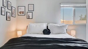 A neatly made bed with white linens and a black throw, a window with a view, and framed pictures on the wall. - Luxury 4BR 2BA House with Pool Spa Billiard Only 6 minutes to the Strip (Las Vegas)