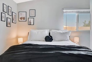 A neatly made bed with white linens and a black throw, a window with a view, and framed pictures on the wall.
