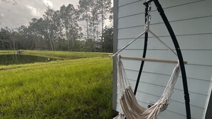 A hammock chair attached to a porch railing, overlooking a grassy field and trees.