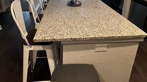 A kitchen with a granite countertop, white chairs, a black trash bin, and a ceiling fan.