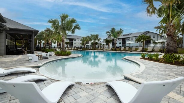 A swimming pool with lounge chairs, palm trees, and a building in the background.