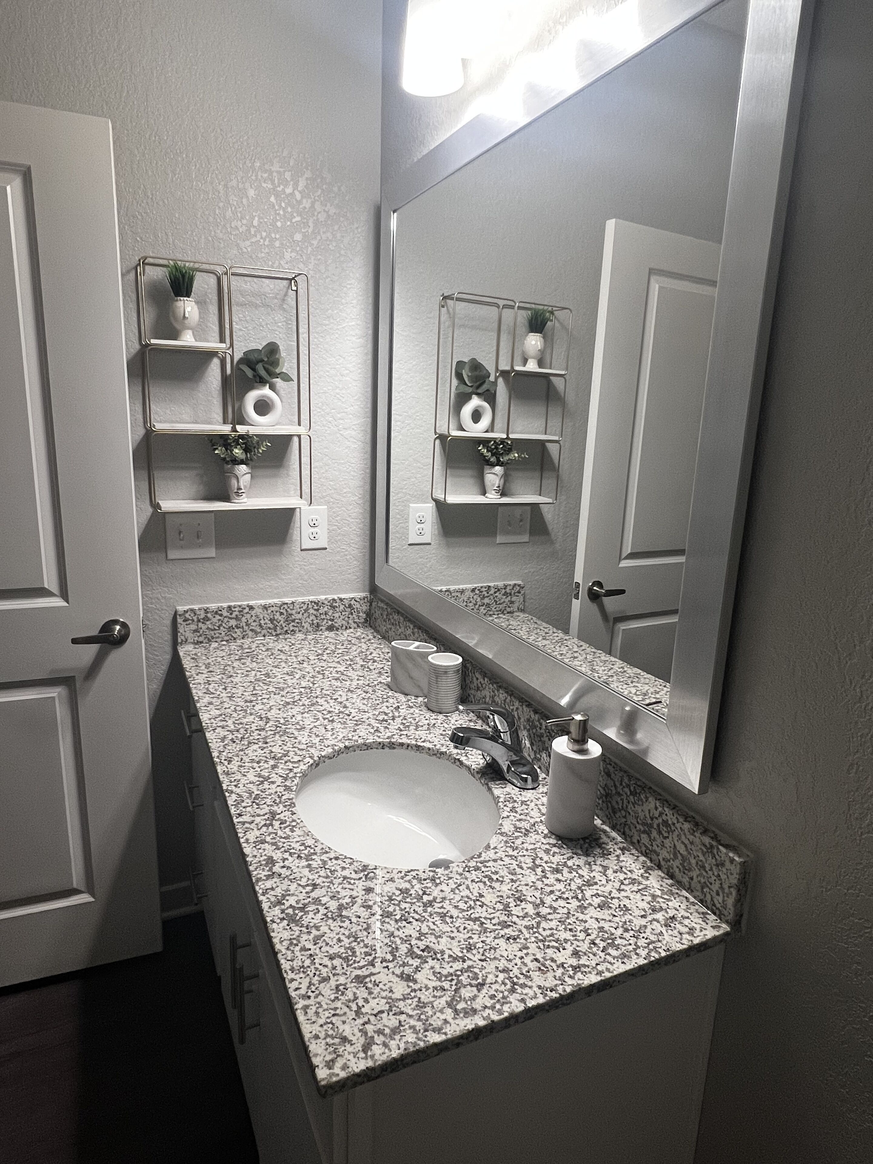 Un baño con encimera de granito, lavabo, espejo y estante metálico con artículos de aseo.