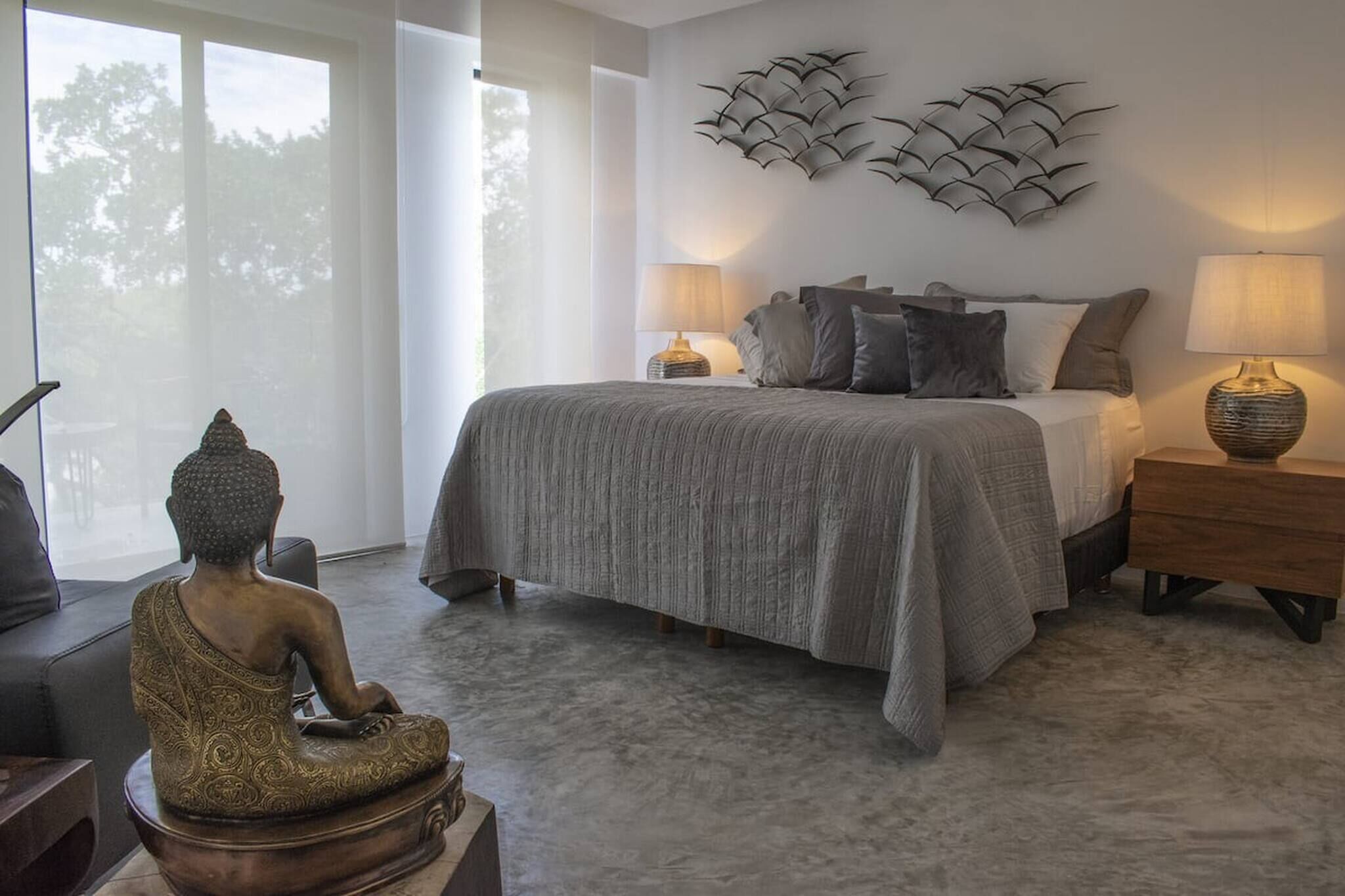 A bedroom with a bed, two bedside lamps, a decorative wall piece, and a Buddha statue.