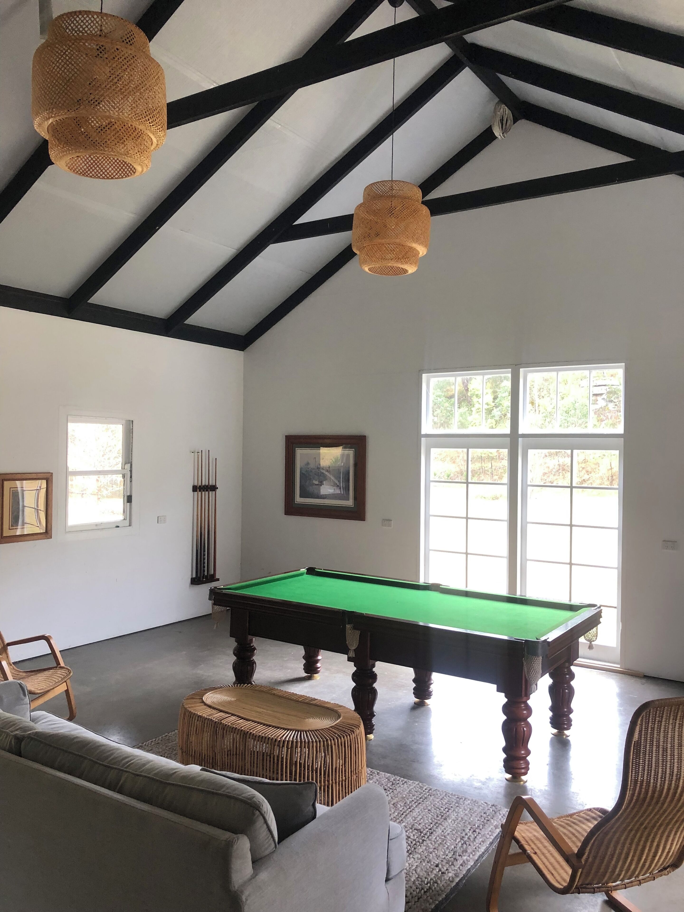 A billiards room with a pool table, two wicker chairs, a sofa, and a large window with natural light.