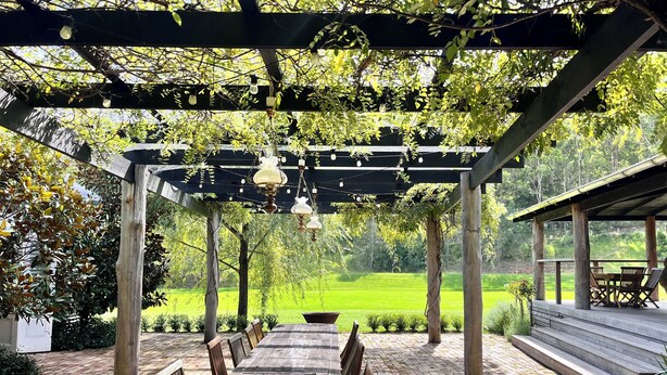 Une terrasse extérieure avec une table et des chaises en bois, une pergola agrémentée de guirlandes lumineuses et une vue sur un champ verdoyant.