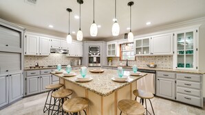 Una cocina amplia con armarios blancos, una isla de granito con taburetes y luminarias colgantes.