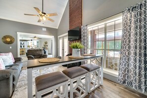 A spacious living area with a high ceiling fan, a dining table with stools, a sofa, and a large window with patterned curtains. - Broken Bow Vacation Rental w/ Pool & Hot Tub! (Broken Bow)