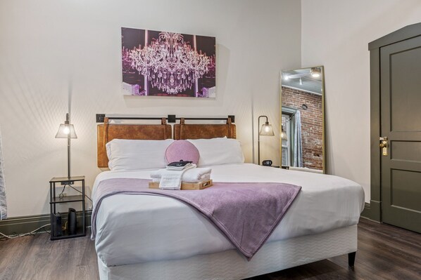 A neatly made bed with a white and purple blanket, a wooden headboard, and a pink pillow. There is a small bedside table with a lamp and a speaker. A large mirror reflects a brick wall and a door. - Historic Main Street 1BR Near Dining and Nightlife (Saint Charles)