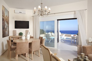 Ein modernes Wohnzimmer mit Essbereich, einem Fernseher und einer großen Schiebeglastür, die auf einen Balkon mit Pool und Meerblick führt.