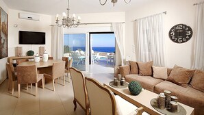 Ein modernes Wohnzimmer mit einer Couch, Esstisch, Stühlen, einer Deckenleuchte, einem Fernseher und einem großen Fenster mit Meerblick.