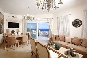 A modern living room with a sofa, dining table, chairs, a chandelier, a TV, and a large window with a view of the sea.