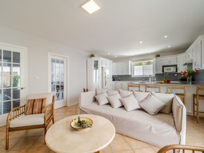 A modern living room with a beige sofa, a round coffee table, and wicker chairs. A kitchen with white cabinets and a dining area with a wooden table and chairs is visible in the background. - Villa Kostamaria, spacious 5 Bed, 3Ba, Sea view, private pool (petriti)